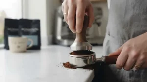 A wooden espresso tamper being used to level fresh coffee grounds in a basket.
